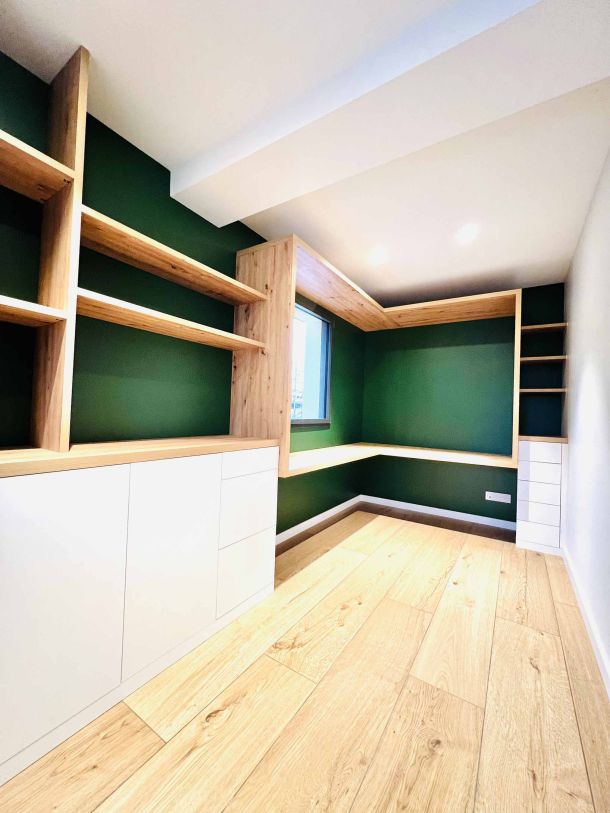 Bureau sur mesure avec étagères et rangements en bois clair, combinés avec des éléments blancs et un mur vert profond, dans un espace moderne au sol en parquet clair et éclairage encastré.